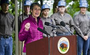 A woman speaks at an outdoor podium while young people in hard hats look on. 