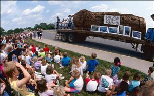 A large group of children sit on the grass looking at a large log on a flatbed truck. The log has a banner that says Save the Ancient Forests. On the back of the truck a man is addressing the crowd and news cameras are pointed at him