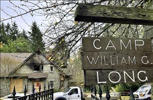 A wooden sign with the text Camp William G Long in the foreground. A one-story building boarded up building with signs of a fire in the background