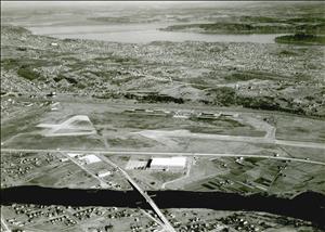 Birds eye view of a single runway with industrial buildings and a river in the foreground and residential neighborhoods around a large lake in the background. 