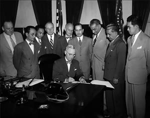 Nine men in suits stand around a seated man who is signing something on his desk. 