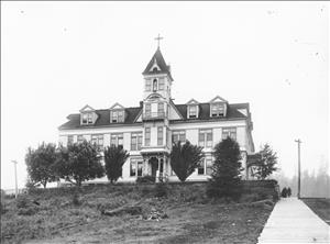 A three story building with a large tower in the middle and peaked windows on the top floor. There is a cross on top of the tower. The building sits at the top of a grassy hill behind a row of small trees. 