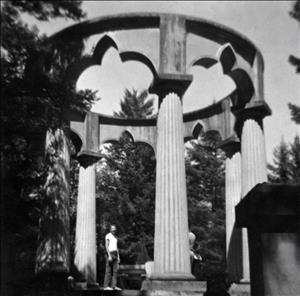 Black and white photo of pavilion surrounded by circle of columns holding an arched ring, with man in white t-shirt standing next to one column 