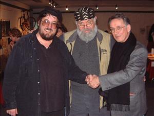 Dorpat wearing a patterned hat with his arms around Stein and Crowley as they shake hands in front of him