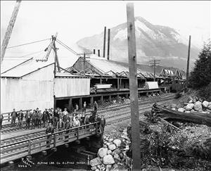 Many men in work clothes stand on railroad tracks in front of large wooden factory buildings. In the background, a large, snow covered mountain peak. 