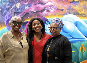 Three smiling Black women with a colorful painting behind them