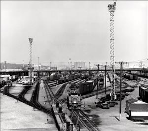 Many railroad tracks pass under a roadway and merge. Telephone poles and radio towers stand between the tracks. Trucks and campers are parked alongside the tracks. 