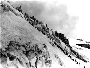 A long line of people in a row ascending a steep snowy slope. 