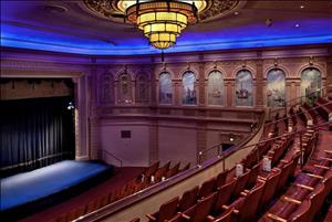 Looking down from a theatre balcony onto a stage with a black curtain. A yellow art deco chandelier hangs from the ceiling and the wall is lined with panels painted to depict old venice. 