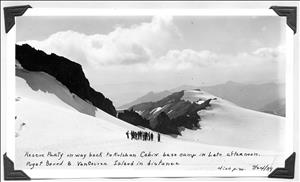 A group of people stand in a cluster in the middle of a snow covered field beneath peaks and crags of a mountaintop. Beneath the photo, handwritten text says rescue party on way back to kulshan cabin base camp in late afternoon. 