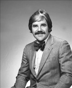 Head and shoulders portrait of a white man with shoulder length hair and a large mustache. He is wearing a suit jacket and bowtie. 
