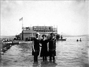 Three people in early twentieth century swimwear stand arm in arm knee deep in water at the beach. Behind them is a wooden boathouse with people sitting along its edge. Canoes are tied to the boathouse. 