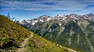 A trail hugs the side of a steep hill covered in low growing vegetation. The bluff looks out on snow capped peaks and a blue sky. 