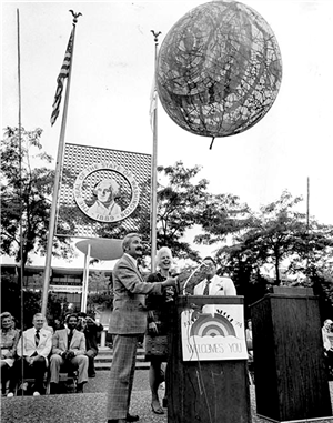 Three people at an outdoor podium smile as a large balloon floats above them. People in suits are seated behind them. Flagpoles and the Washington State seal are in the background. 