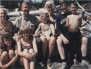 A family of two adults and six children sit close together on a log on a beach. The sun is shining and everyone is smiling. The youngest child is sitting on the woman's lap. 