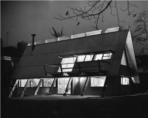 An a frame house at night. All of the lights are on inside the house. Windows along the slanting roof are illuminated. 