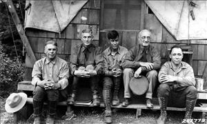 Five men sit in front of a wooden structure with a canvas roof. They are wearing uniforms similar to those of forest rangers with hats and boots. 