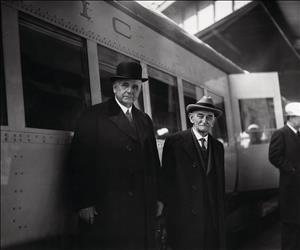 Two elderly men wearing hats, ties, and long winter coats, stand in front of a train car. 