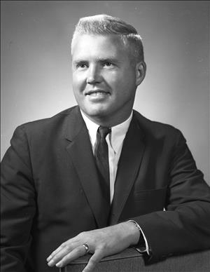 Head and shoulders portrait of a white man with blond hair wearing a suit and tie. He is resting his left hand on the arm of a chair. 