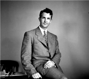 A young white man in a suit and tie sits on a desk. 