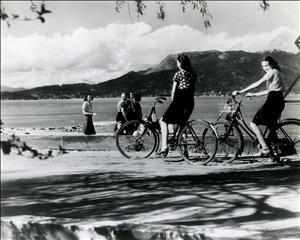 Two people sit on their bikes on a paved path along the shore of a lake. Other people stand on the beach, In the distance clouds push up against a hillside. 