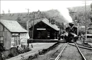 A steam locomotive is stopped in front of a small wooden building with a sign on it that says Colfax. There are people standing beside the train. A smaller wooden building next door has an advertisement painted on the side that says Golden West Coffee Just Right. 