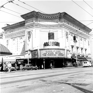 A large white building on the corner of a city block. It has a decorative overhang along the top of the building and a large arched entry way with a sign that says Colisuem. The marquee says winner of three academy awards steve mcqueen and paul newman towering inferno. 