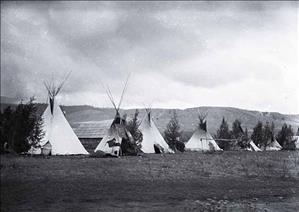 Black and white photo of line of teepees in front of trees at edge of open field, with hills rising beyond