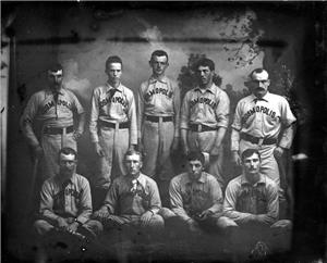 Nine people in baseball uniforms pose for the camera. Five of them are standing behind the other four who are kneeling. 