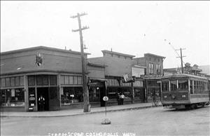 A small town intersection with a row of one story storefronts. A trolley approaches the intersection. 