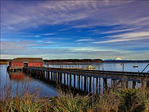 A long wharf extends out to a large wooden structure Penn Cove. In the distance is Camano Island, and beyond that a snow-capped mountain in the Cascade Range