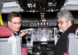 Two white men sitting in the cockpit of an airplane look back over the shoulders at the camera