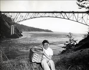 A child sits with a picnic basket under their arm and a big smile on their face. Behind them is a tall steel bridge spanning a narrow waterway. 