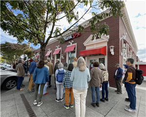 A large group of people with their backs to the camera face a one story brick building on a city sidewalk. 