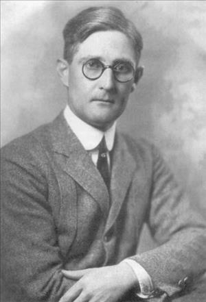 Black and white head-and-shoulders photo portrait of a man in jacket and tie wearing glasses.