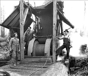 Three men stand around a large piece of machinery with a spool in the middle and cable running off of the spool. 