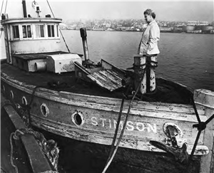 A woman in a raincoat stands on the deck of an old wooden boat tied to a pier. She has her hands in her pockets and is looking down at the deck of the boat. On the bow of the boat is painted the word Stimson. In the distance the shoreline of the lake is lined with houses and industrial buildings. 