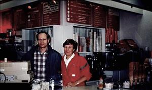 Two people stand behind a cash register and beneath menu signs. 