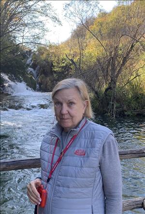 A woman stands on a bridge over a stream against a background of yellow and green trees. 