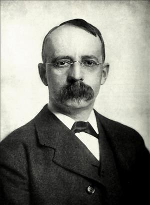 A head and shoulders portrait of a man in a suit and cravat with rimless eyeglasses and a very large mustache. 