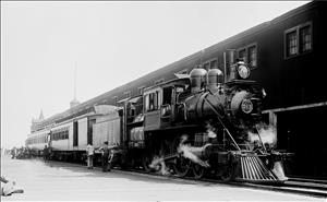 A steam locomotive with one passenger car behind it is stopped in front of a two story building. Men in conductor uniforms stand beside the train. 