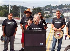 An elderly indigenous woman speaks at a podium, in the middle of a road, with six indigenous people behind her