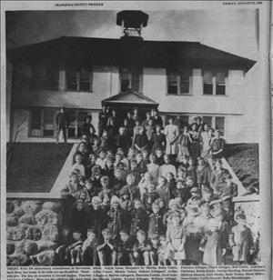 Portion of newspaper page with black and white photo of large group of children posing on the steps of a two-story schoolhouse