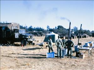 Men in military uniform stand around an outdoor kitchen. Behind them trucks and canvas tents on a grassy field