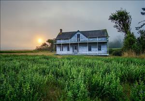Photo of Ferry House, an old, white, wood-frame building on the uplands of Ebey's Landing on Whidbey Island. The house is in the distance, and to its left is either a rising or setting sun. Ferry House has been at that location since 1860 and in now part of Ebey's Landing National Historical Reserve.