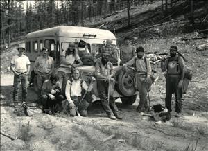 A group of people in jeans and work shirts stand around, and sit on, a short boxy bus, in a clearing in the forest. They are holding tools and wearing backpacks. There are two dogs, one laying at a person's feet and another in the arms of someone who is kneeling. 