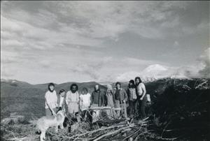 Nine people in work clothes with bushy beards and long hair stand in a clear cut on top of a mountain. They are laughing and posing for the camera. A snow capped mountain is in the background. A big dog is with the group of people. 