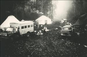 An encampment in a foggy forest. A pickup truck with a large camper on its back, two yurts with smoke coming out of their stove pipes, and a Volkswagen bus are all parked in the clearing. Debris is scattered on the ground. A man with a bushy beard carries gear through the middle of the camp. 