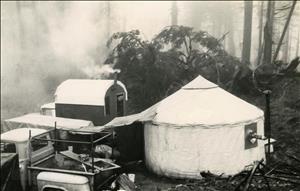 A round canvas tent, a pick up truck, and a makeshift camper in a foggy forest. Smoke rises from a stovepipe on the camper. A person with a beard and winter hat peers out of the camper door. 