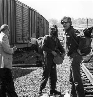 Two men wearing backpacks, one with a bushy beard, stand between railroad tracks and a freight train. A third person, wearing a suit, is writing something down. 